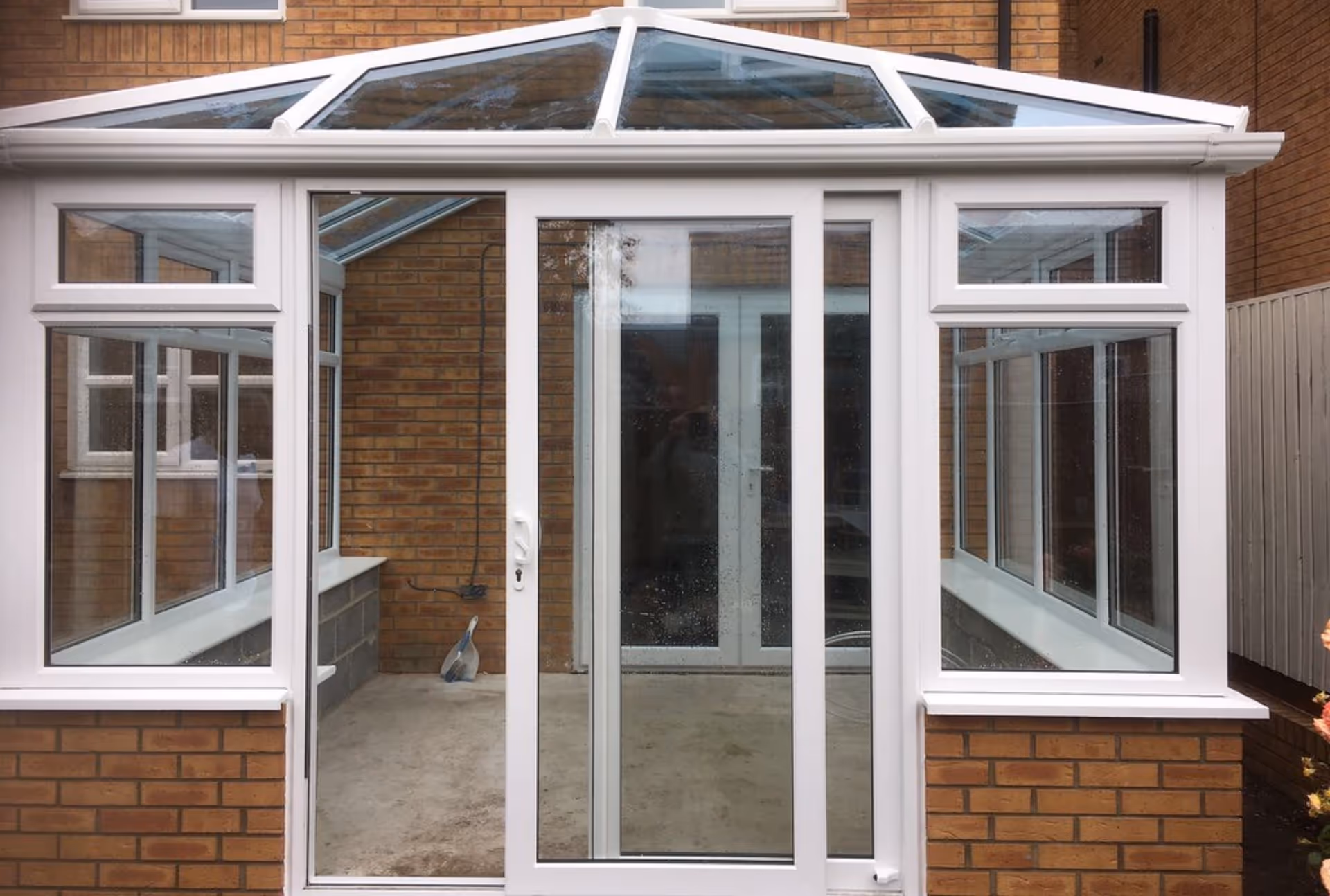 Brick house with white-framed conservatory featuring large glass windows and a central sliding door, with an unfinished interior floor visible.