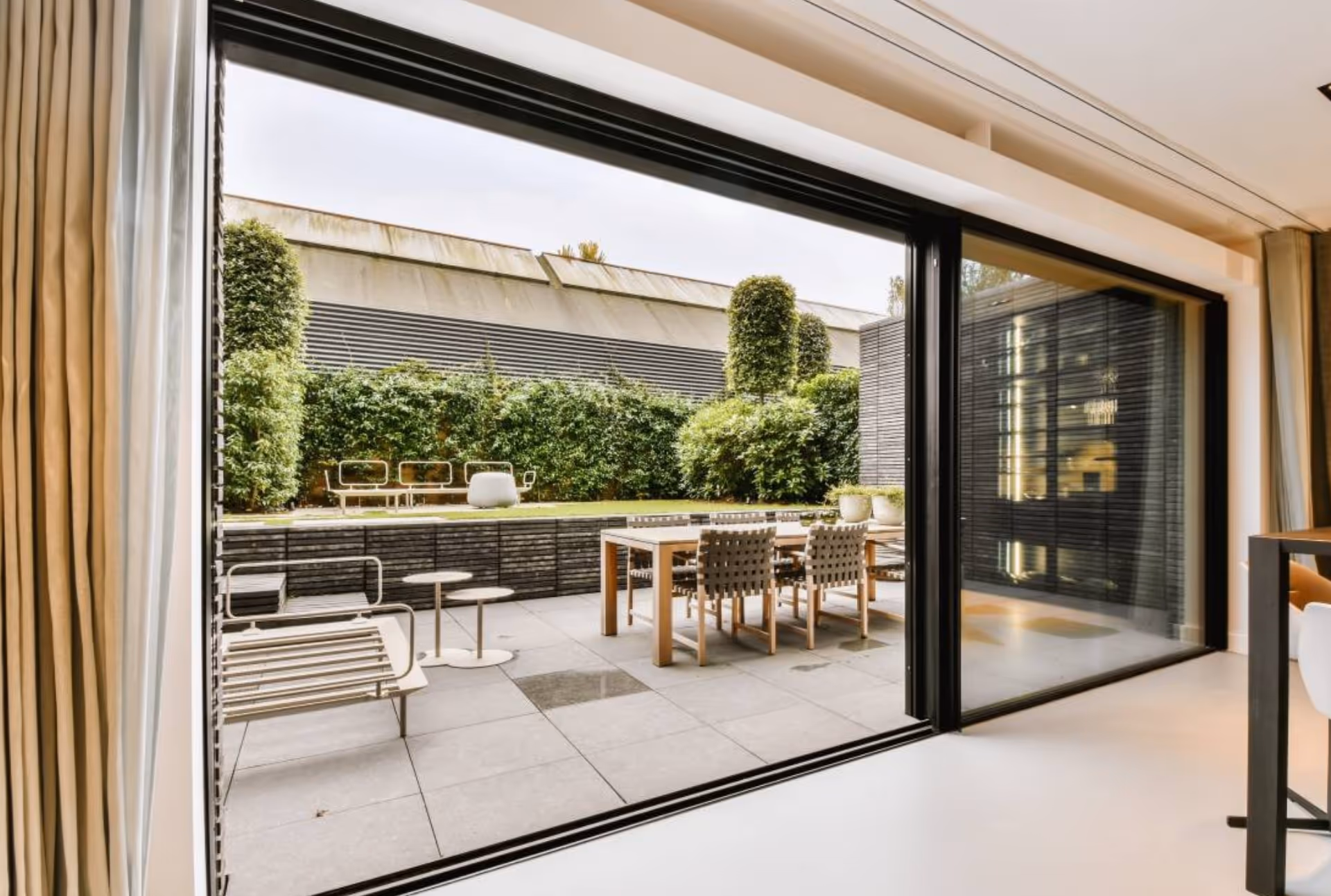 Contemporary patio with outdoor dining furniture, seen through large sliding glass doors, surrounded by manicured hedges and topiary trees.