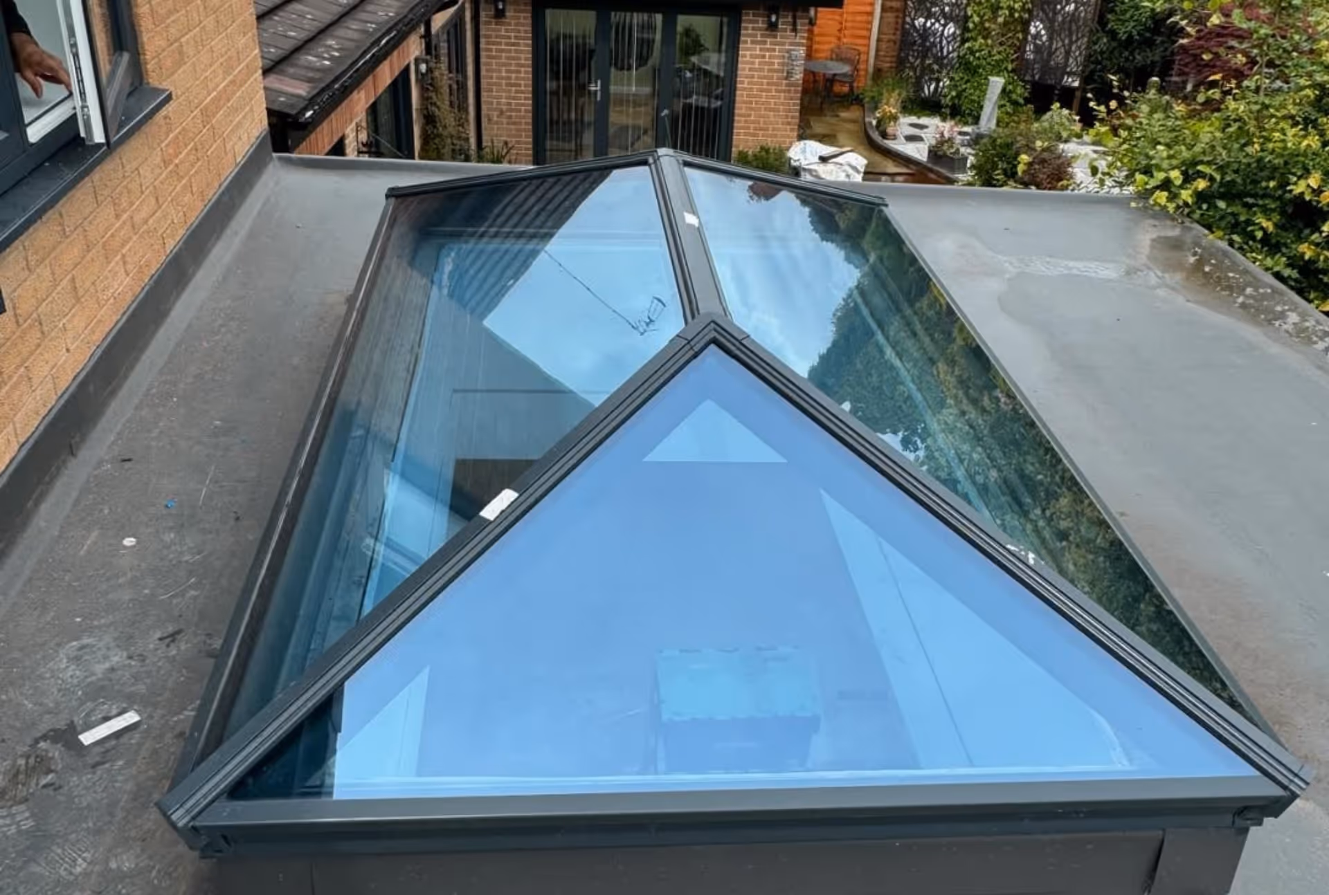 A pyramid-shaped skylight installed on a flat roof, with reflections of the sky and surrounding structures visible in the glass.