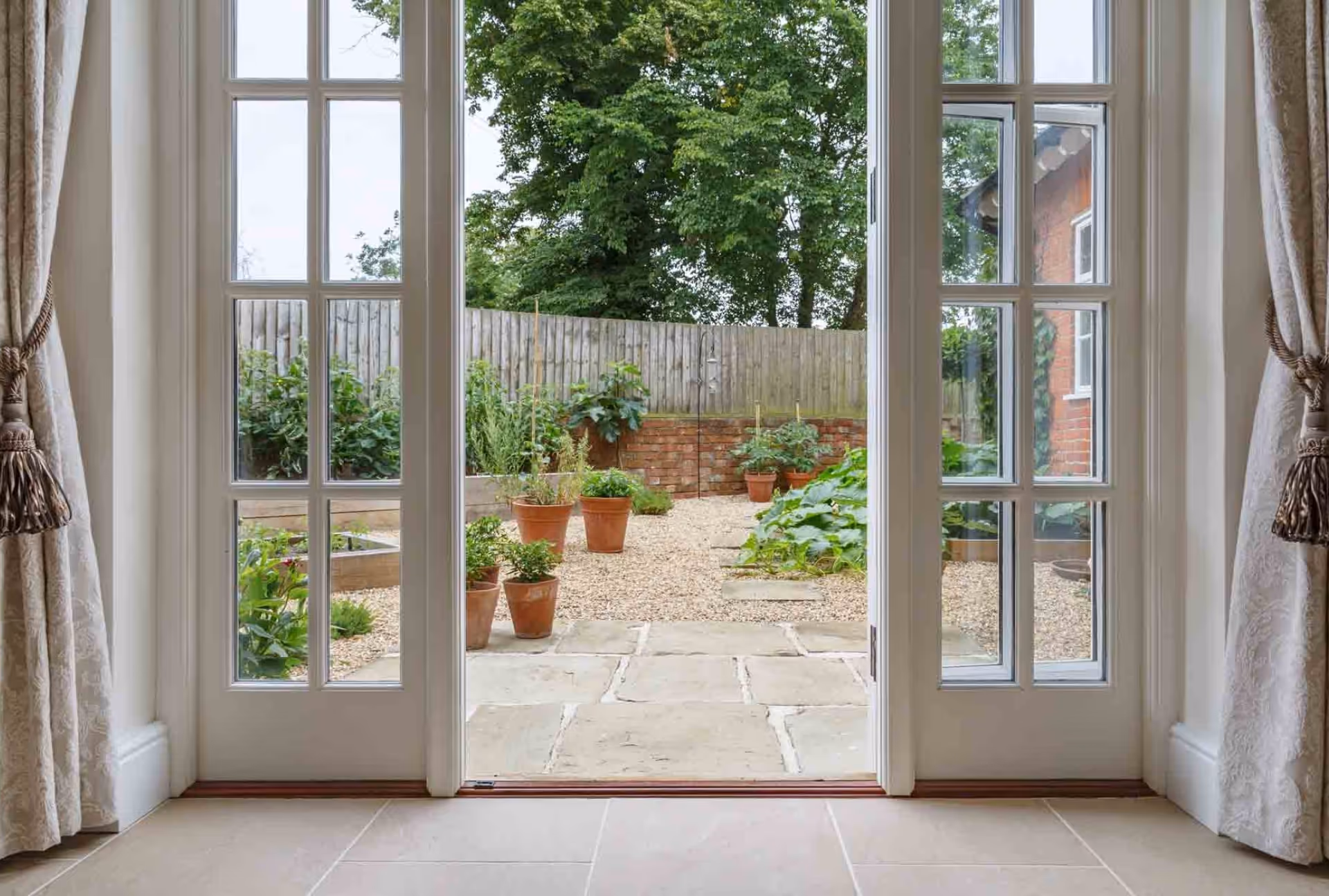 View through French doors onto a well-maintained garden with potted plants, raised beds, and a wooden fence.