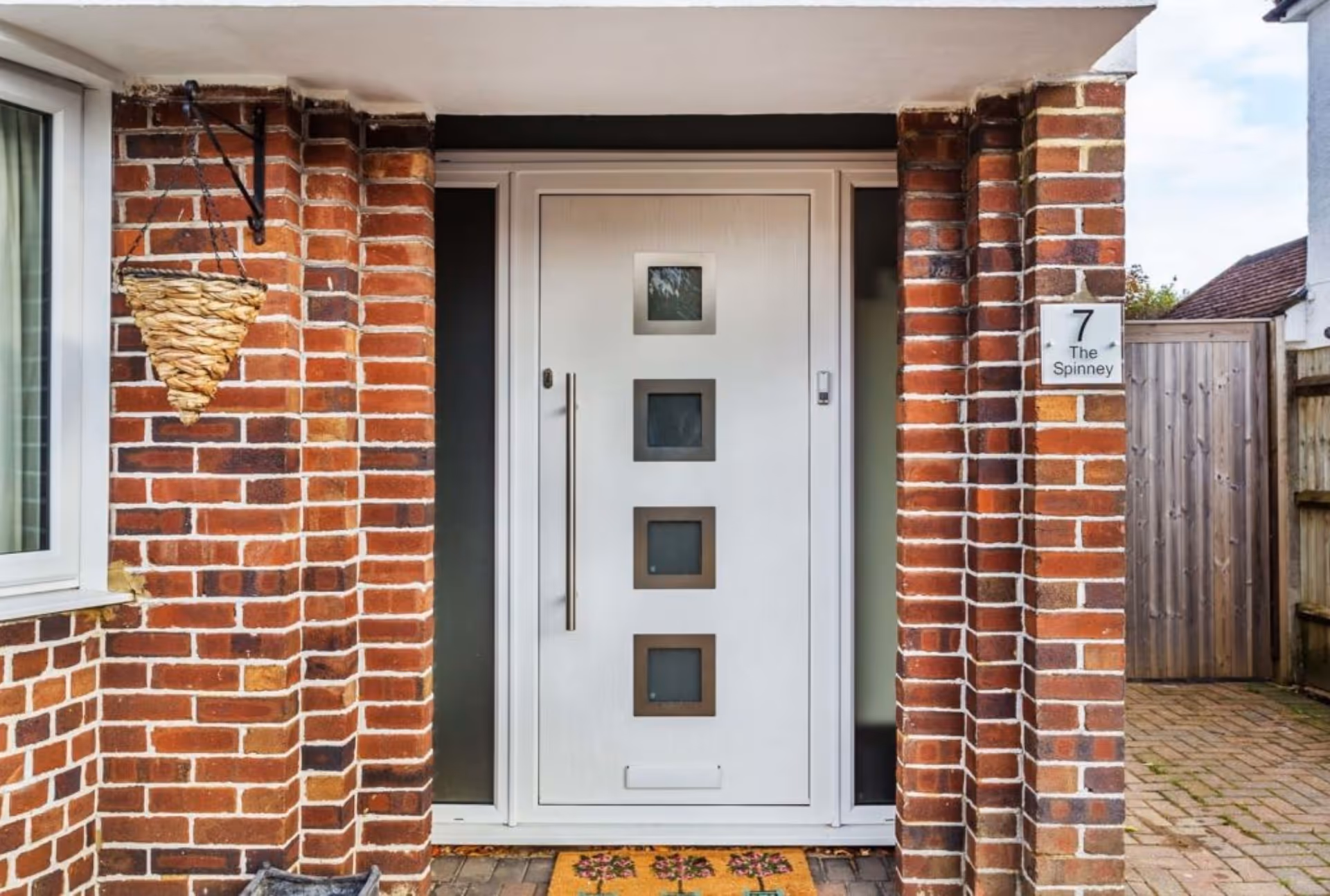 Modern white composite door with three square glass panels, featuring a long metallic handle and set in a brick entryway.