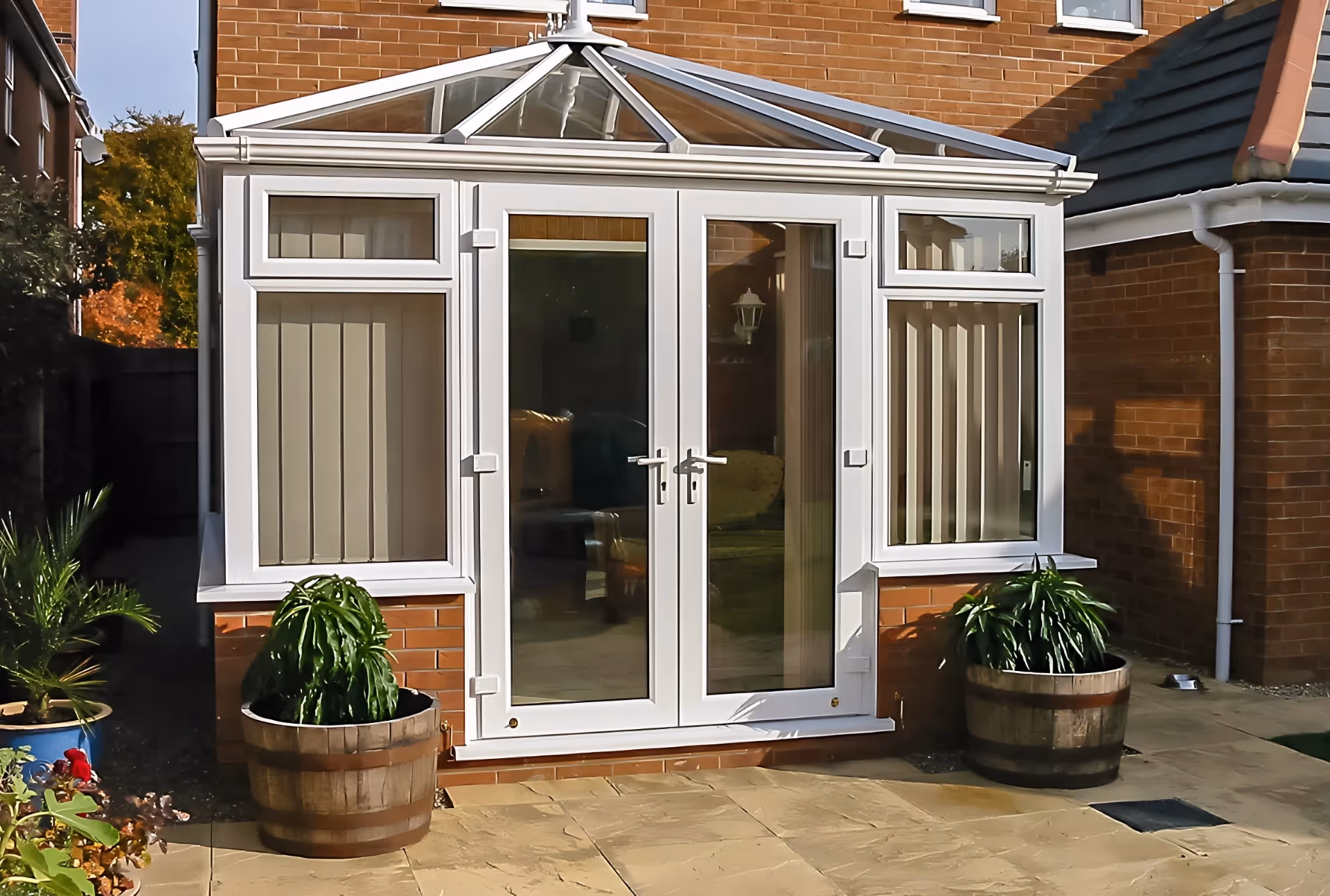 White-framed conservatory with pitched glass roof and central double doors, flanked by large potted plants at the entrance