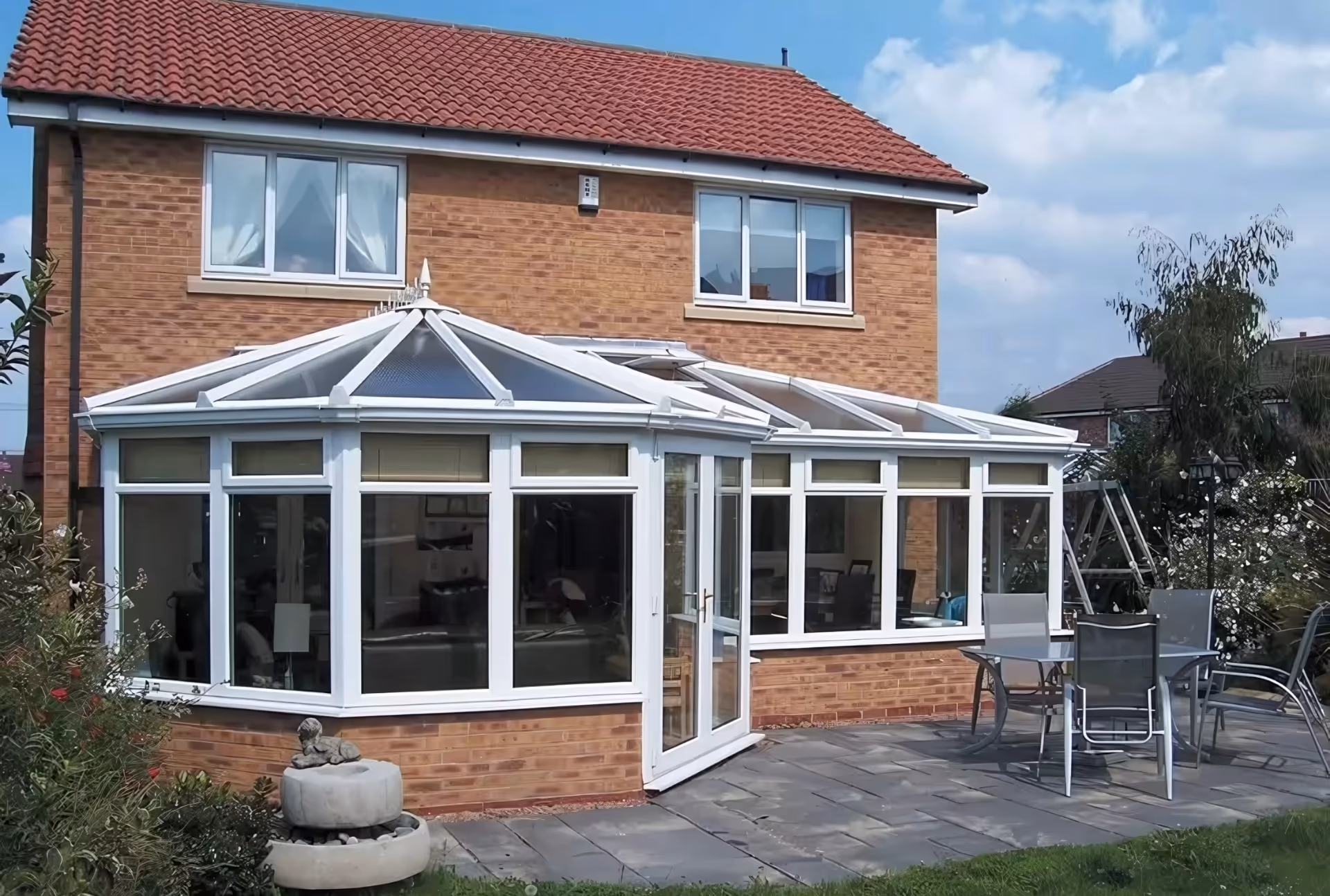 White-framed conservatory with glass windows and doors, attached to a brick house with a red-tiled roof, allowing natural light into the interior.
