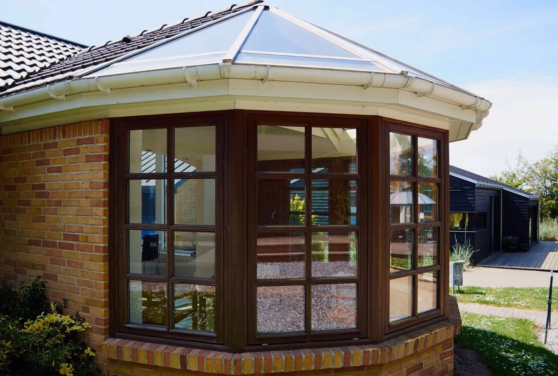 Brown-framed Victorian conservatory with paneled windows and glass roof, attached to a brick house with visible interior furnishings.