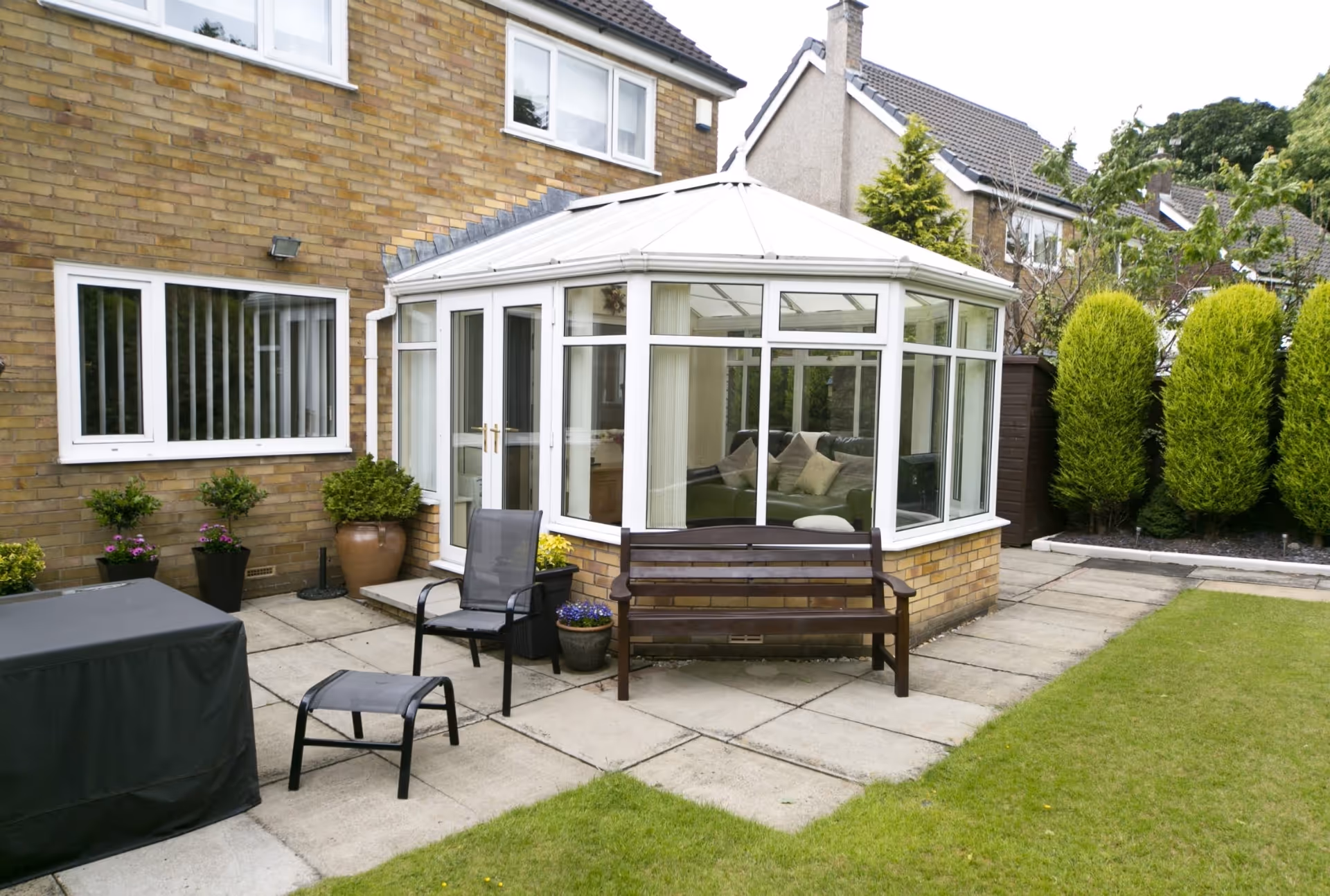 White-framed conservatory with pitched roof attached to a brick house, surrounded by outdoor furniture and potted plants.