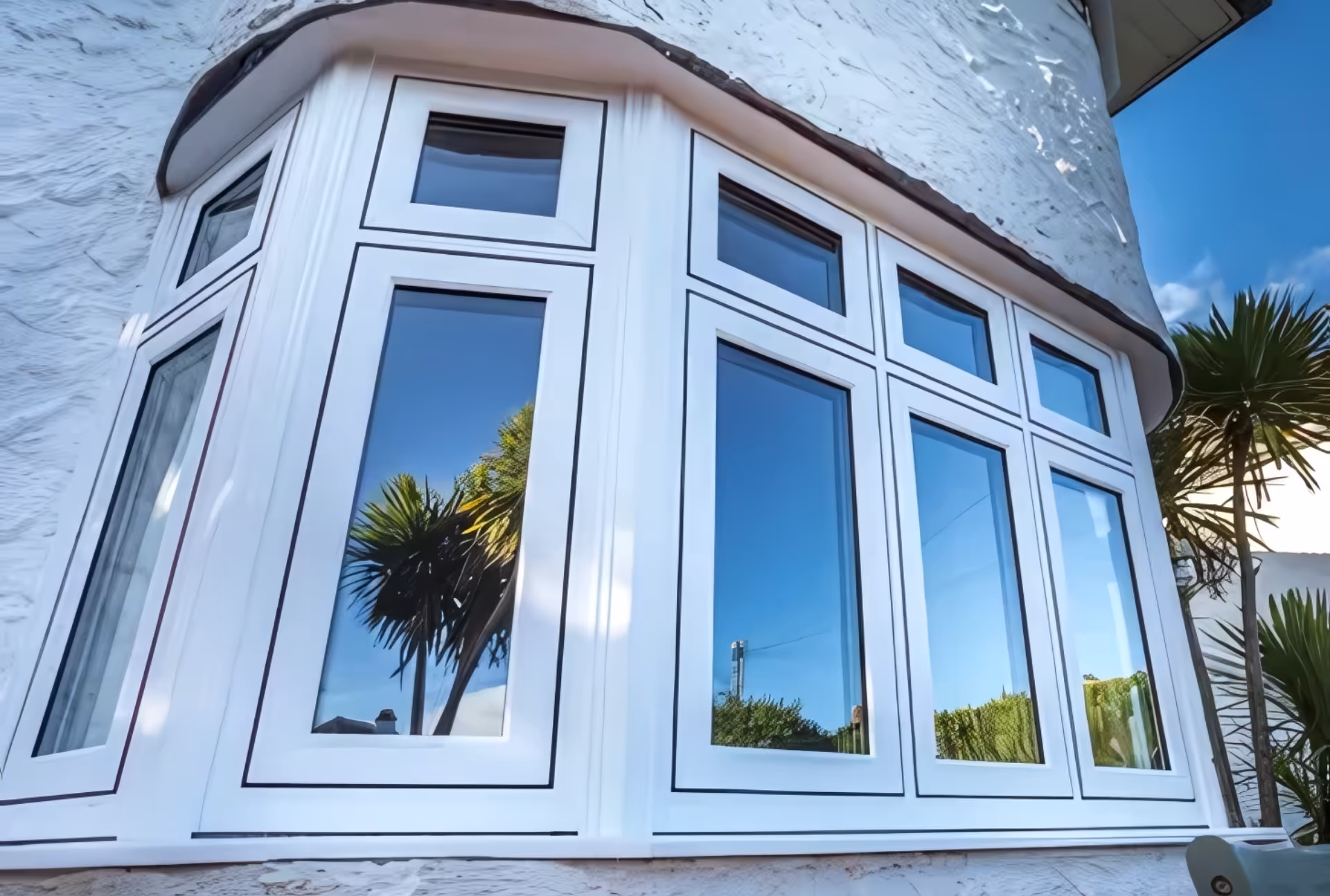 White-framed bay windows projecting outward from a stuccoed wall, reflecting palm trees and clear blue sky.