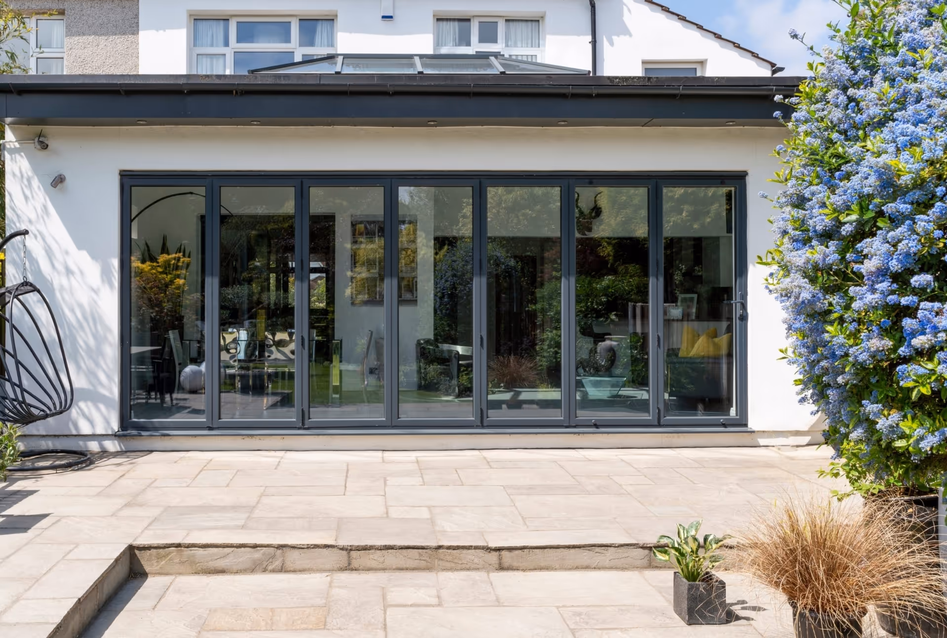 Exterior view of sleek bi-fold doors with large glass panels, framed by flowering bushes and potted plants on a well-maintained stone patio.