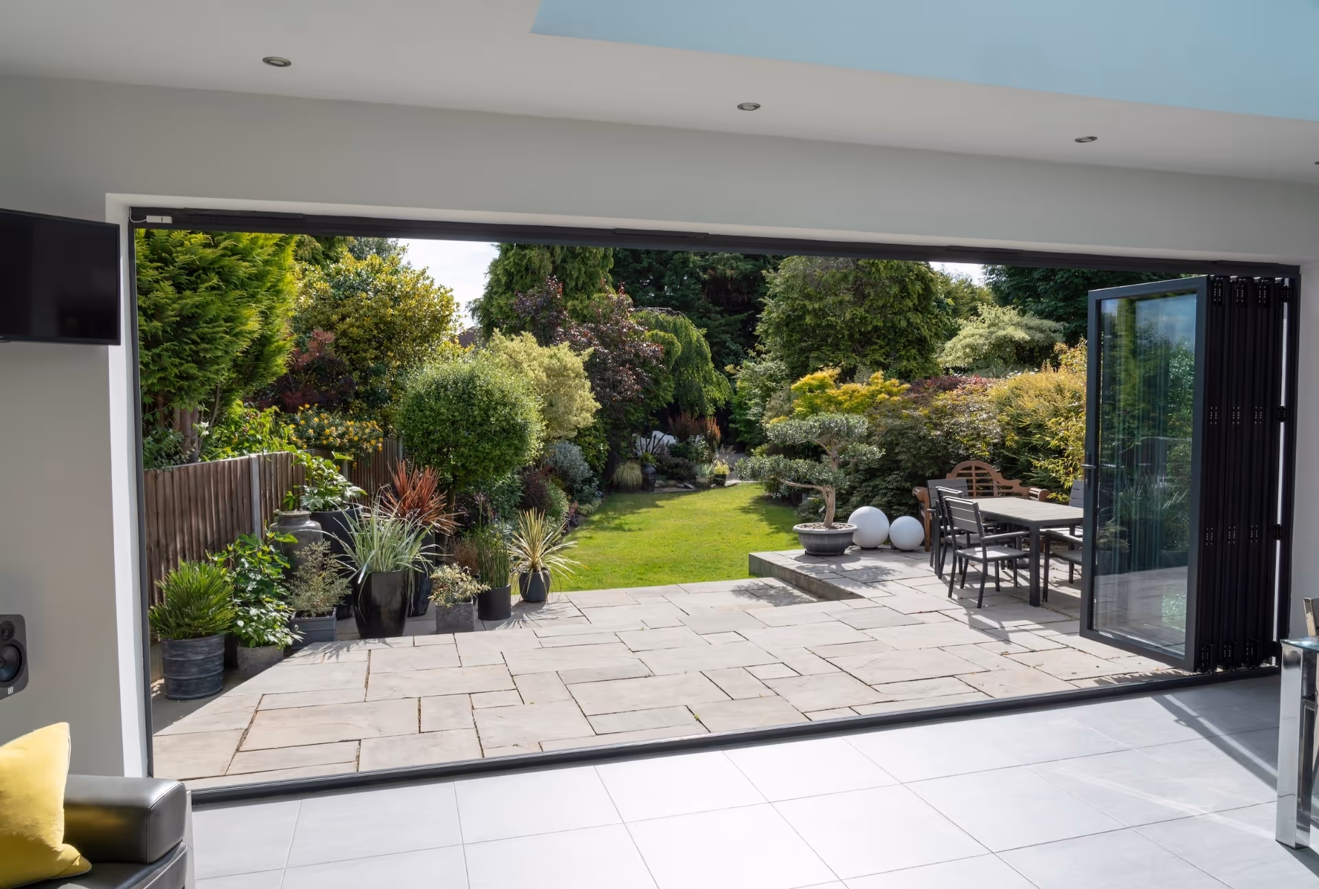 Bi-fold doors fully opened to reveal a stone-paved patio and lush garden, creating a seamless indoor-outdoor living experience.