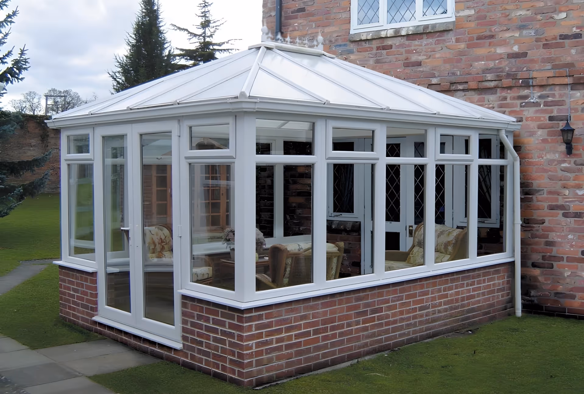 White-framed Edwardian conservatory with pitched roof and clear glass panels, attached to a brick house with visible interior furnishings.