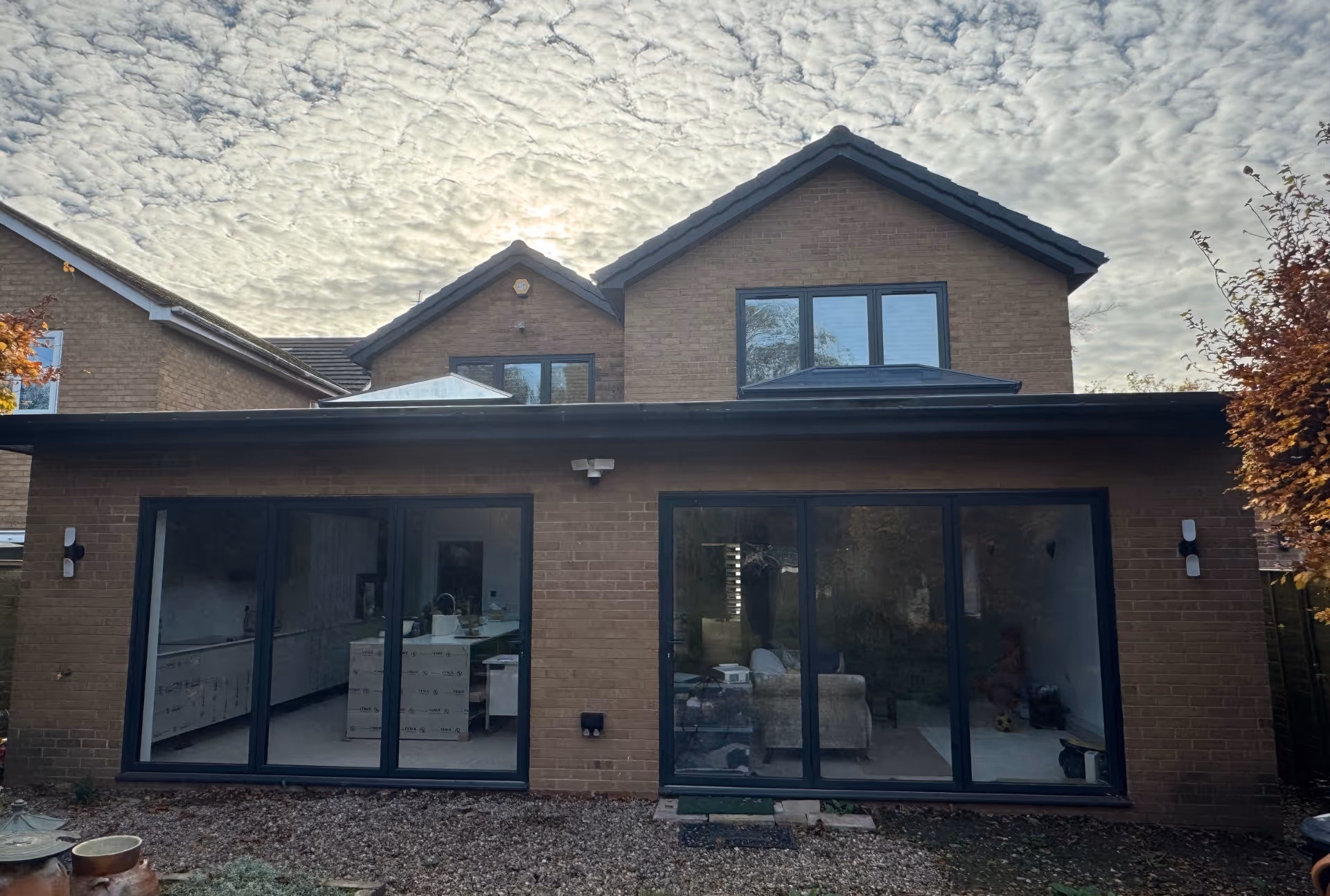 Two-storey brick house with large glass sliding doors opening to an open-plan kitchen and living area, set beneath a sky with patterned clouds.