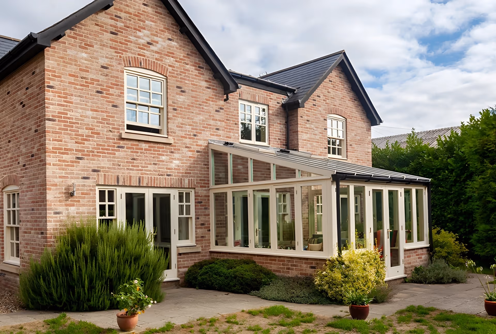 Brick house with an elegant conservatory featuring large windows and a sloped glass roof, surrounded by a well-kept garden with shrubs and potted plants.