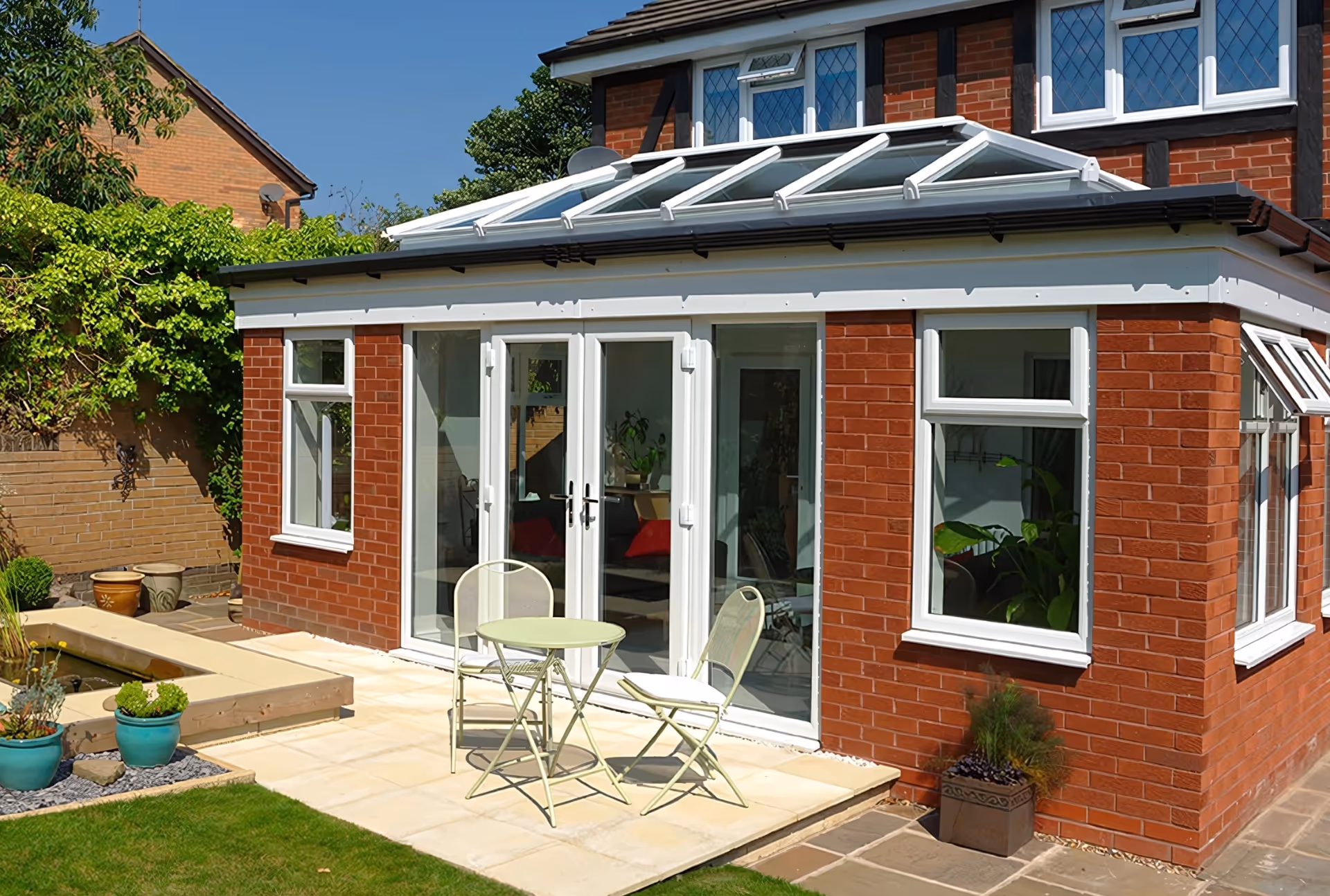 Flat-roof orangery extension with skylights and large glass doors, attached to a brick house with a patio seating area of two chairs and a small table.