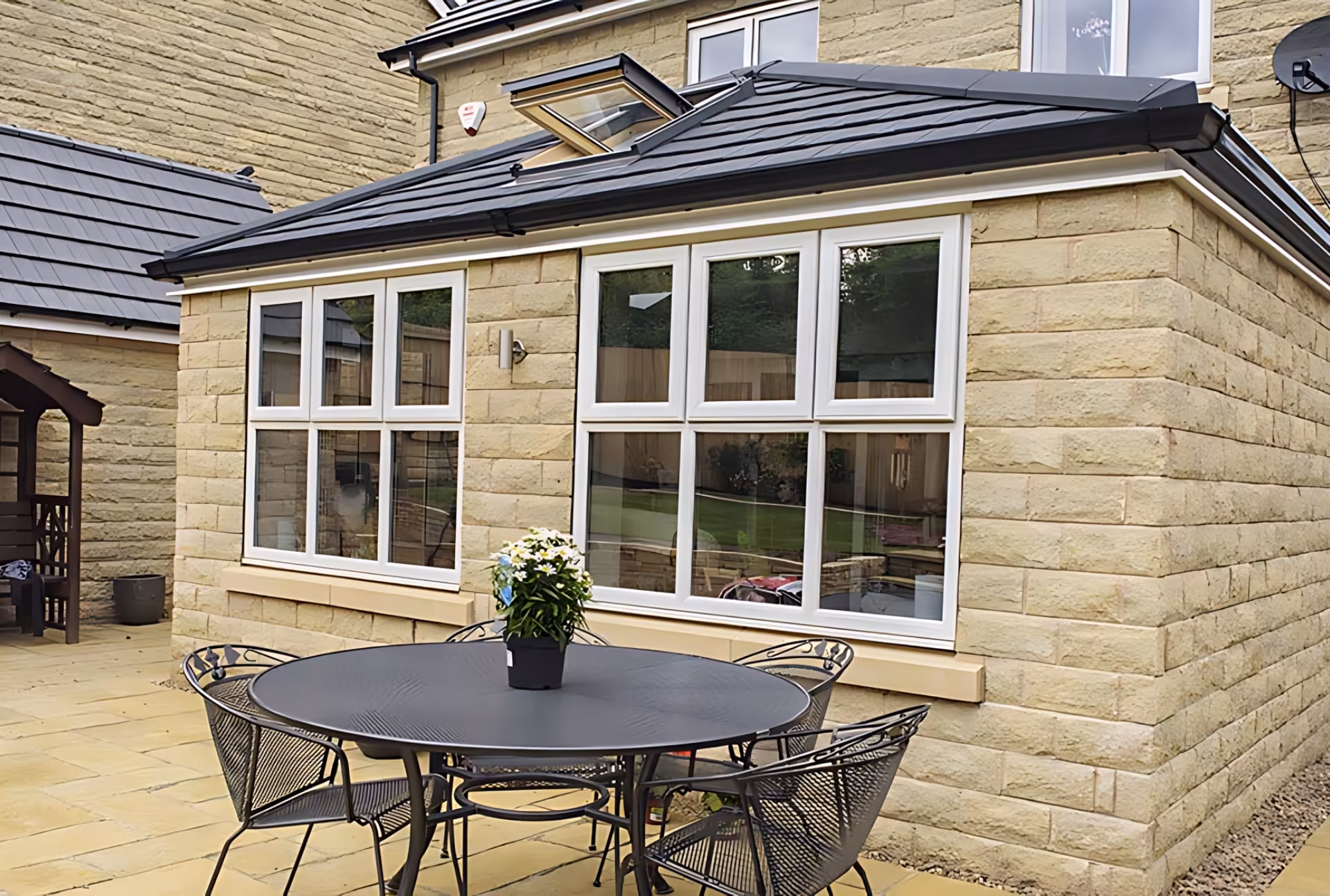 Orangery with white window frames and black tiled roof with skylights, attached to a beige brick house with outdoor seating area.