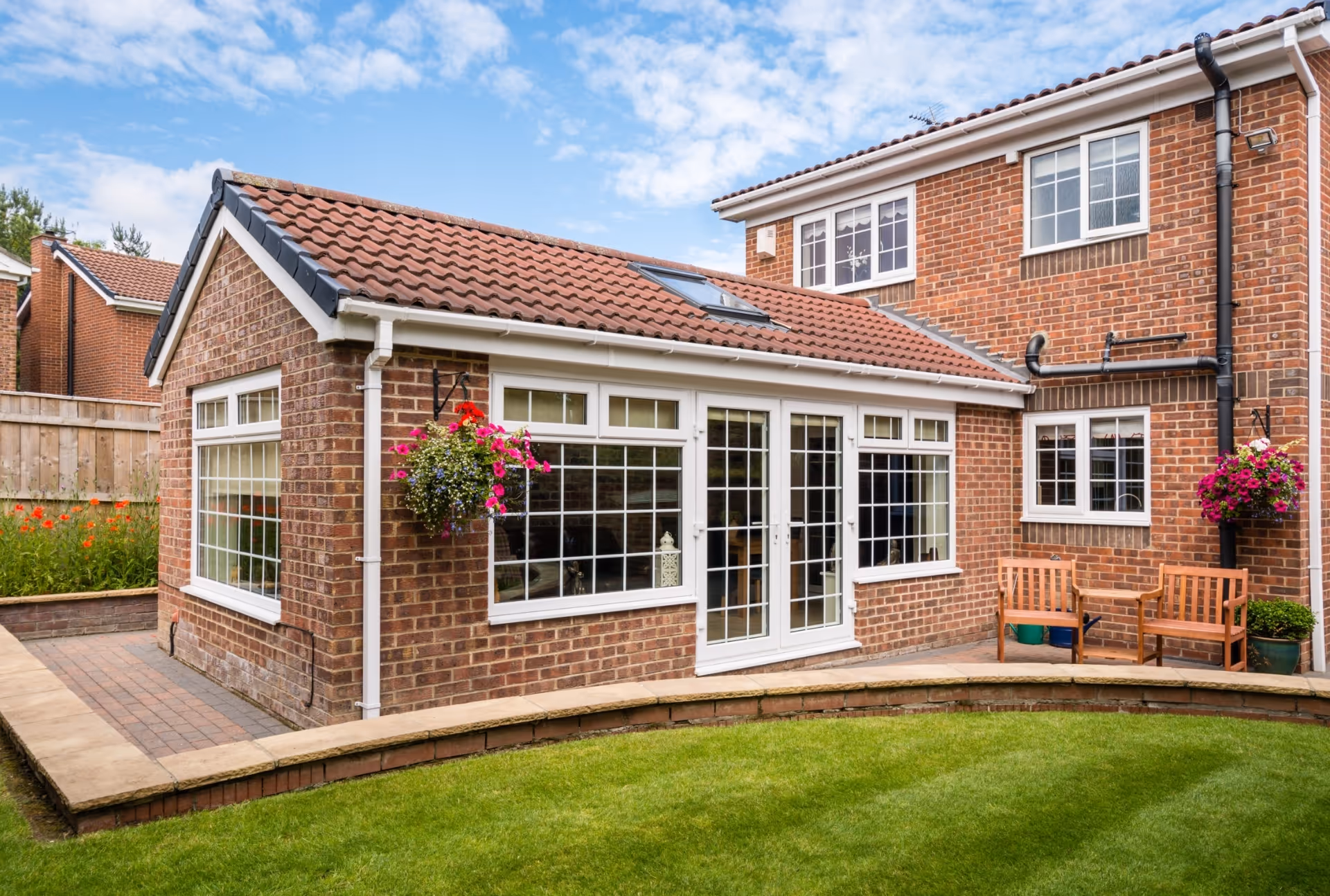 Brick house with a pitched-roof orangery extension featuring skylights, large windows, and hanging flower baskets, overlooking a lawn and paved garden path.