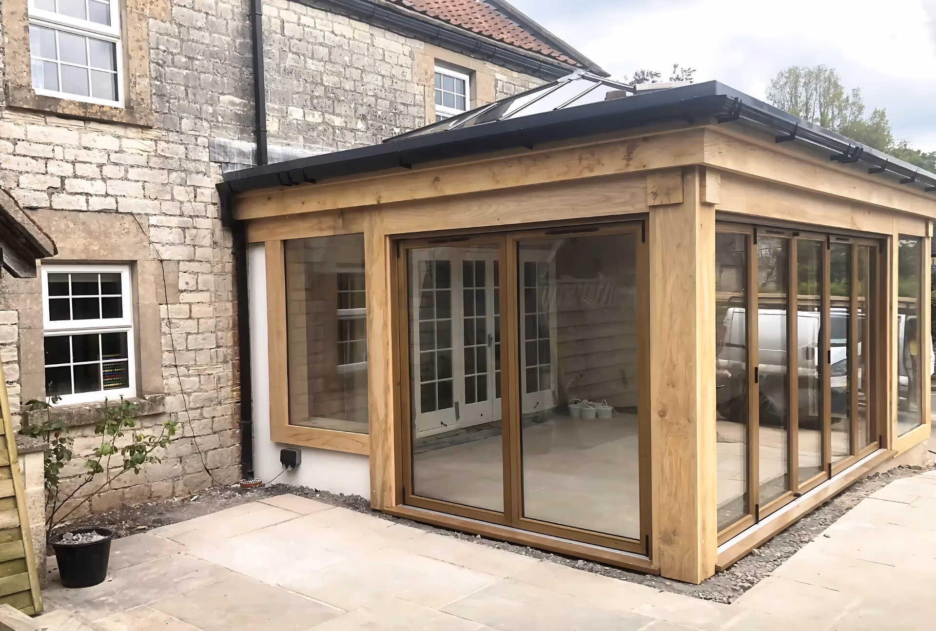 Modern orangery with wooden-framed glass sliding doors and skylights, attached to a stone house.