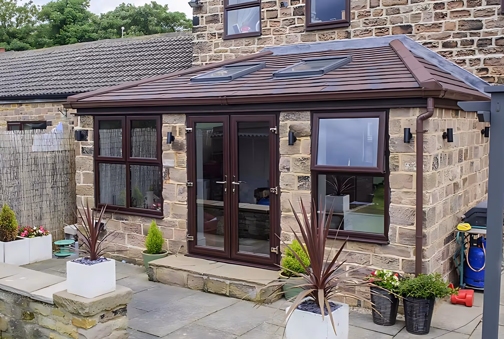 Stone house with a dark-framed orangery featuring double doors, skylights, and surrounding potted plants.