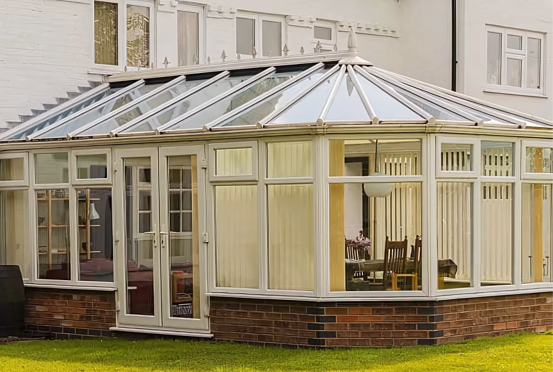 White conservatory with glass roof and vertical blinds, attached to a white house and furnished as an additional living space.