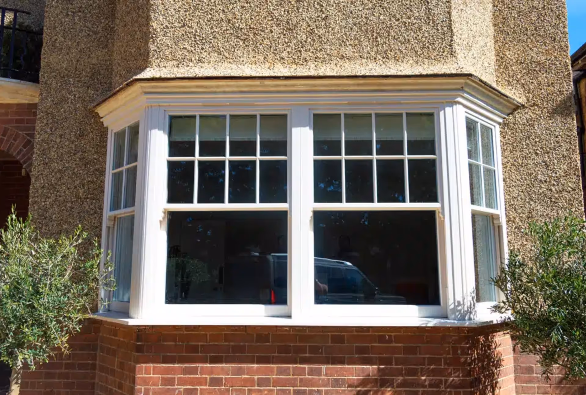 "Large bay window with multiple vertical sliding sash windows, featuring white frames and multi-pane design on a textured and brick exterior.