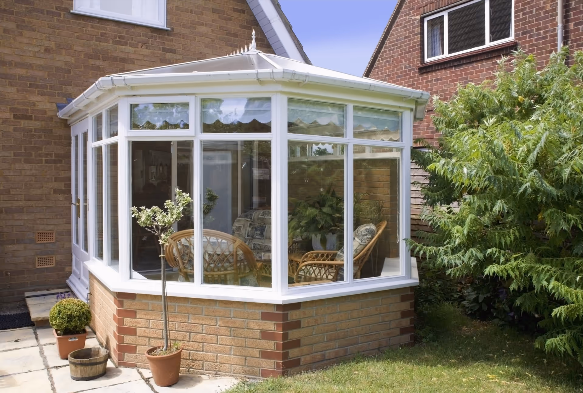 Elegant white Victorian conservatory with large windows and glass roof, featuring wicker furniture and potted plants inside, connected to a brick house.