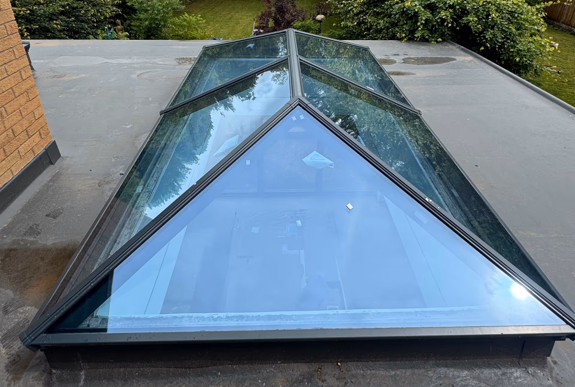 Another view of a large pyramid-shaped skylight on a flat rooftop, capturing reflections of trees and sky.