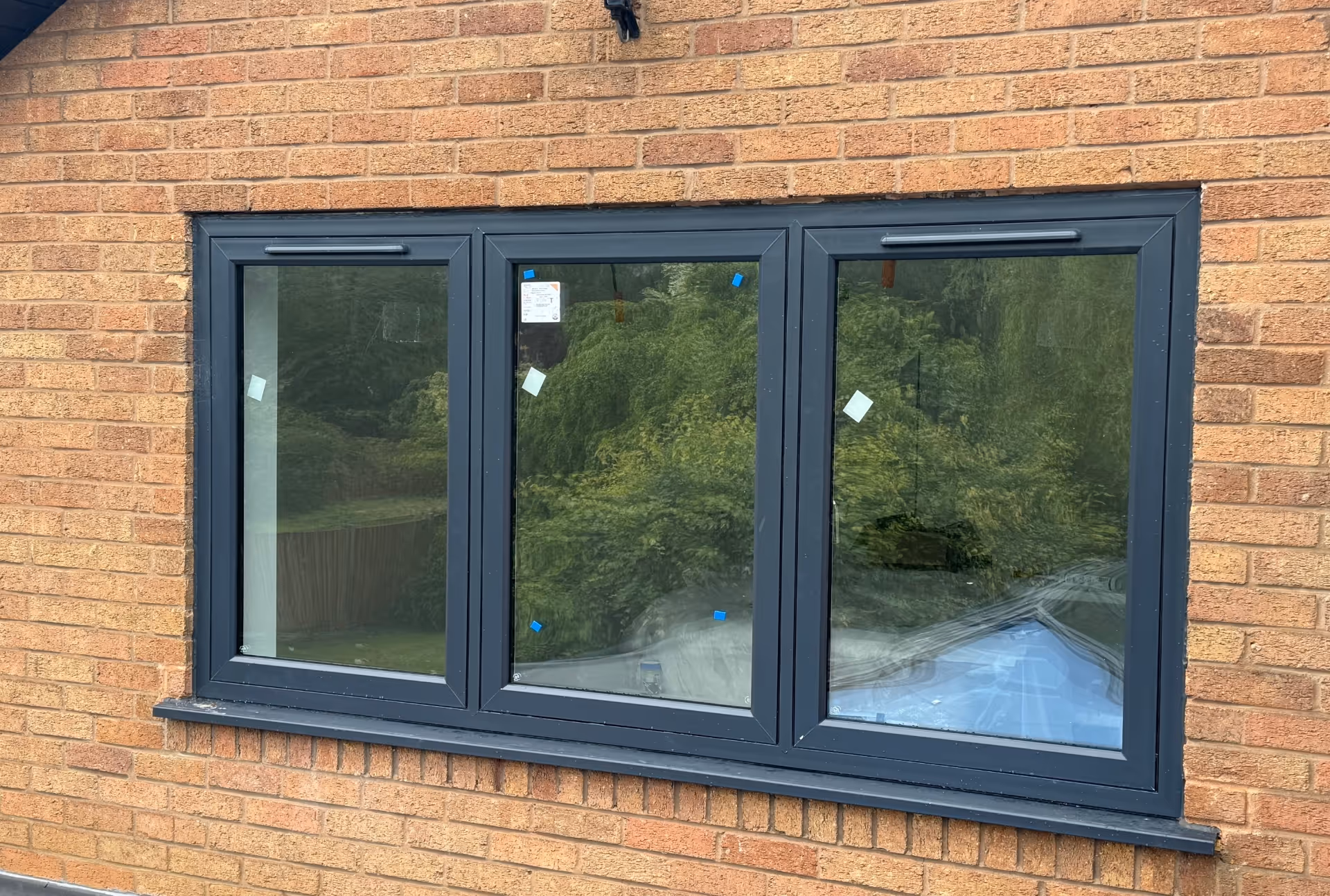 Set of three fixed black-framed windows on a brick wall, reflecting surrounding greenery.