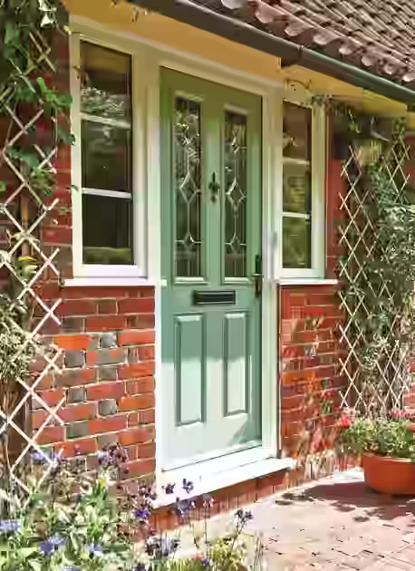 Green front door with glass panels and hardware flanked by white-framed windows on red brick house, by window and door suppliers