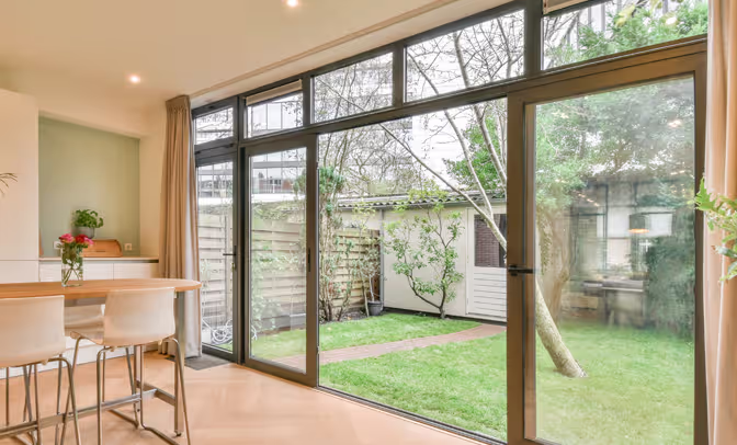 Inviting garden view through sliding glass doors, featuring a tiled patio with wooden table, chairs, and pink flowers, framed by lush greenery.