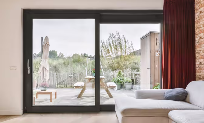 Inviting living room with beige sofa, brick fireplace and bookshelves, viewed through sliding glass doors with potted ferns on terracotta tiles.