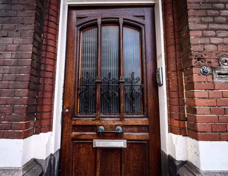 Contemporary dark grey front door with metallic accents and vertical handle, framed by potted plants and a wall-mounted security feature.