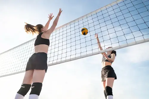 Two female volleyball players jumping near the net with a yellow and black volleyball against a clear sky.