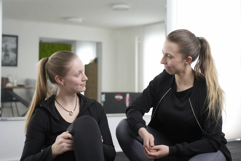 Two young women with ponytails sitting on a floor facing each other, smiling and engaged in conversation in a bright room.