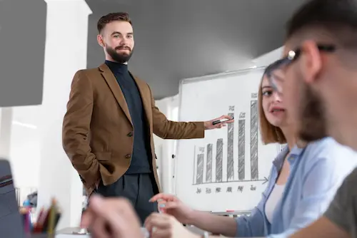 Man in a brown blazer presenting a bar chart to colleagues in a meeting room.