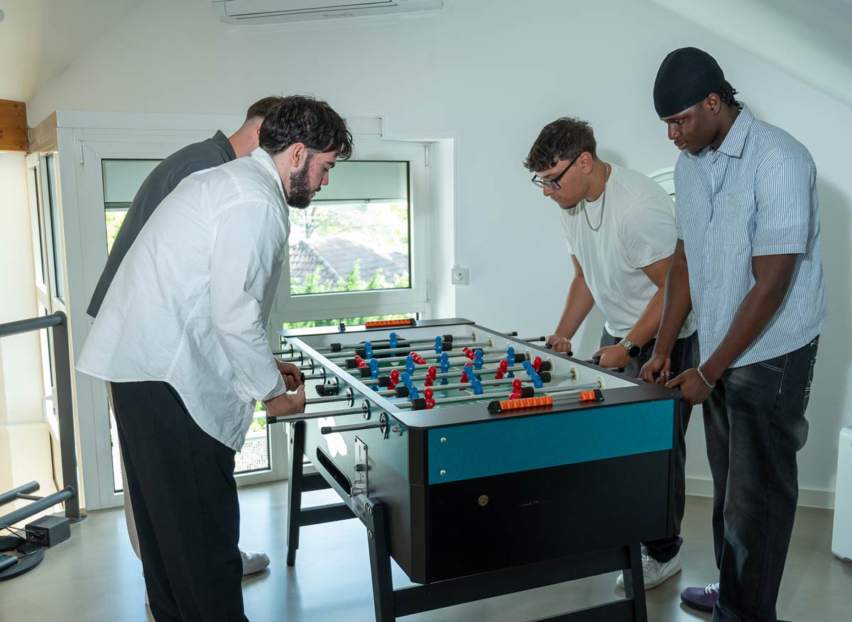 Vier junge Männer spielen Tischfußball in einem hellen Raum mit großen Fenstern und modernem Interieur.