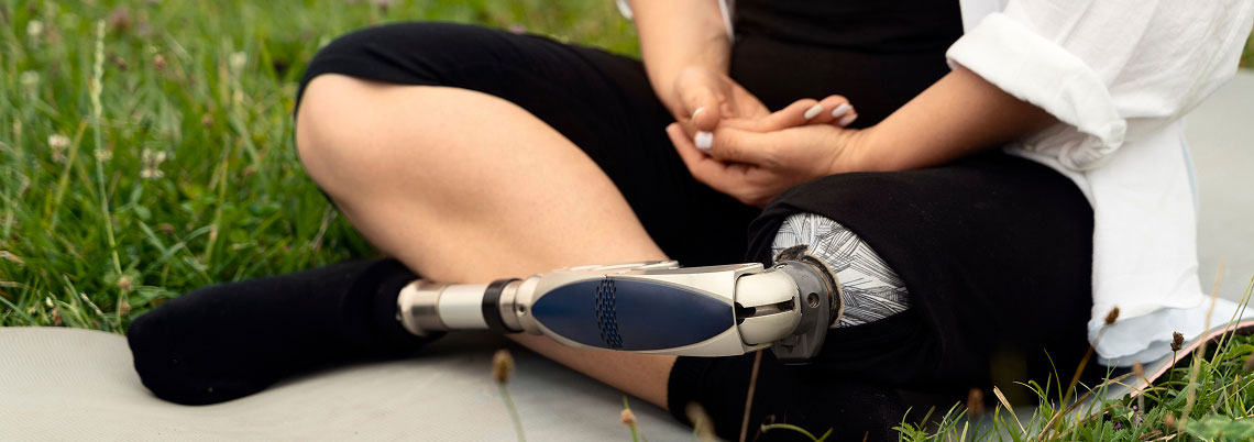 Person sitting cross-legged on grass with a black sock on one foot and a prosthetic leg with a patterned sleeve on the other, hands resting on their knee.