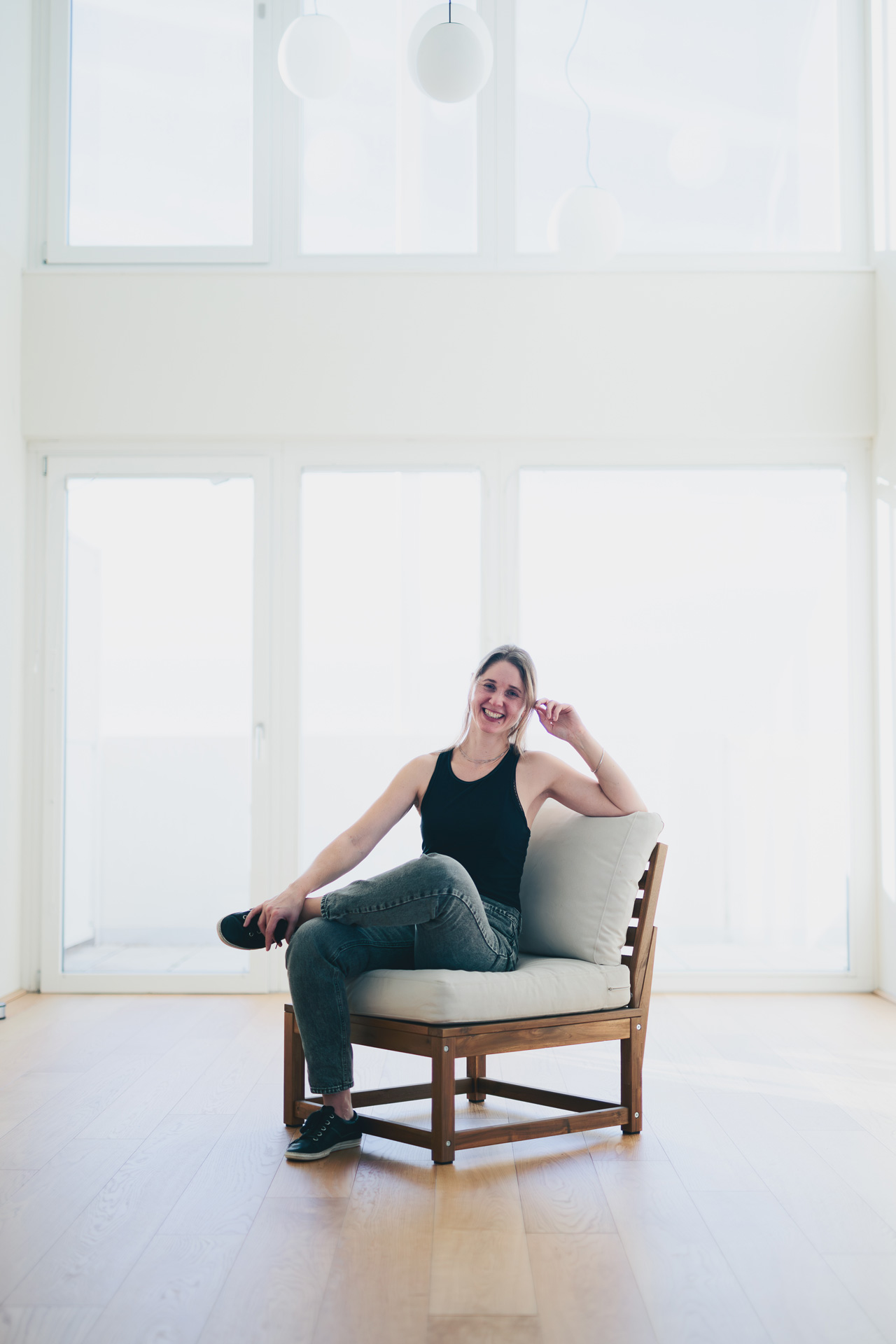 Alexandra sitting on a chair in a bright room with high walls and large windows, smiling to the camera.