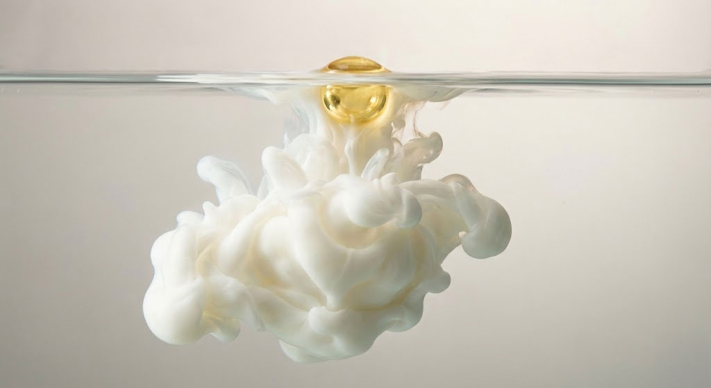 Golden oil droplet suspended at the waterline, releasing a soft milky cloud below—an abstract visual of emulsion, texture, and skincare sensoriality.
