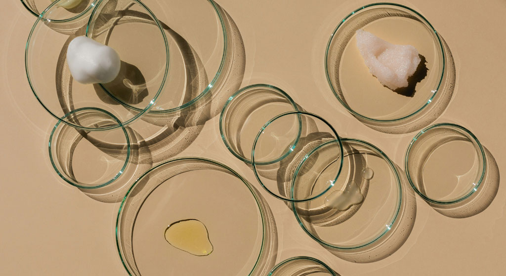 Top-down view of glass Petri dishes on a beige surface, holding small samples of an oil phase and a white material, evoking lab analysis and quality control.