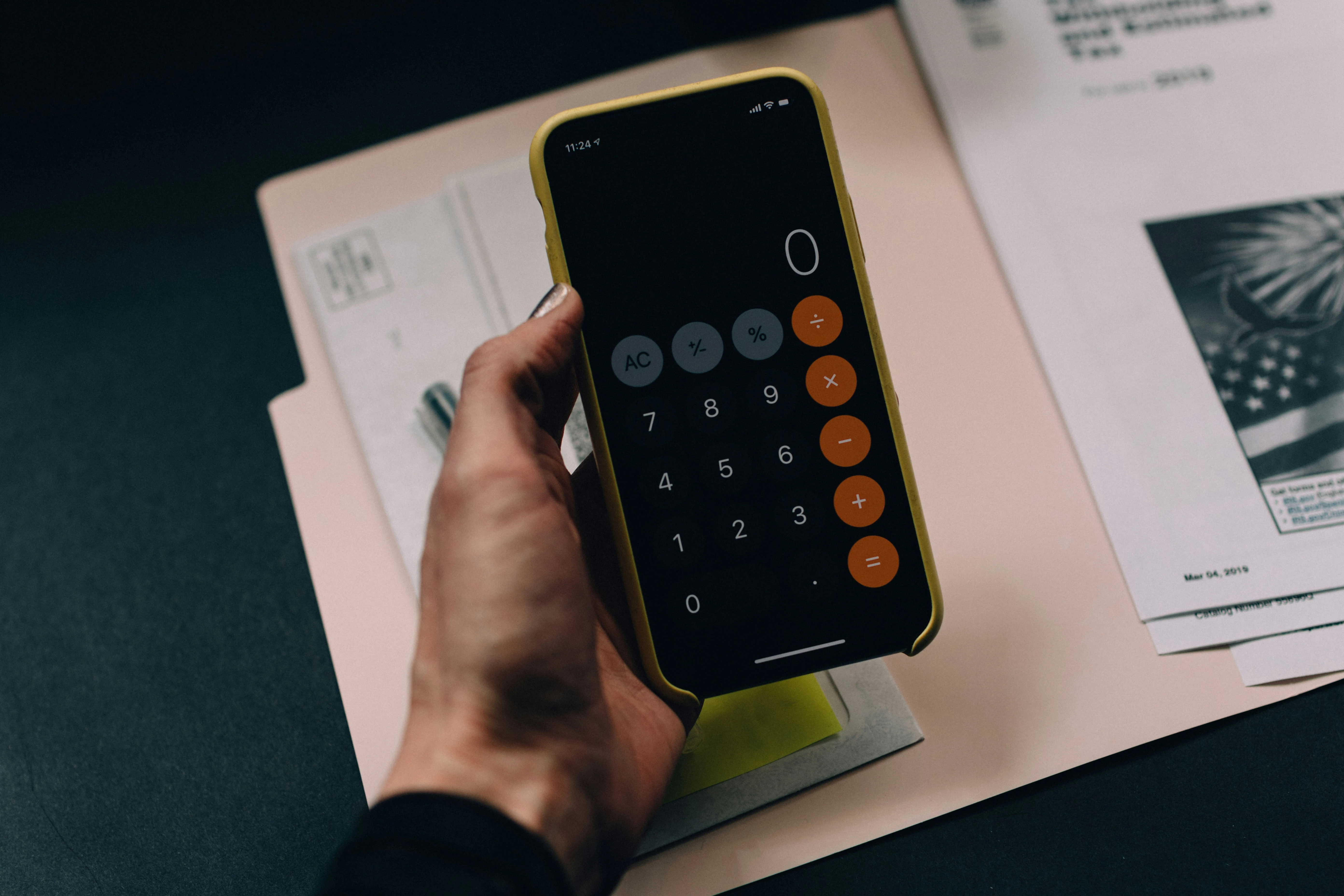 A hand holding a smartphone with a calculator app open, with invoices and documents in the background.