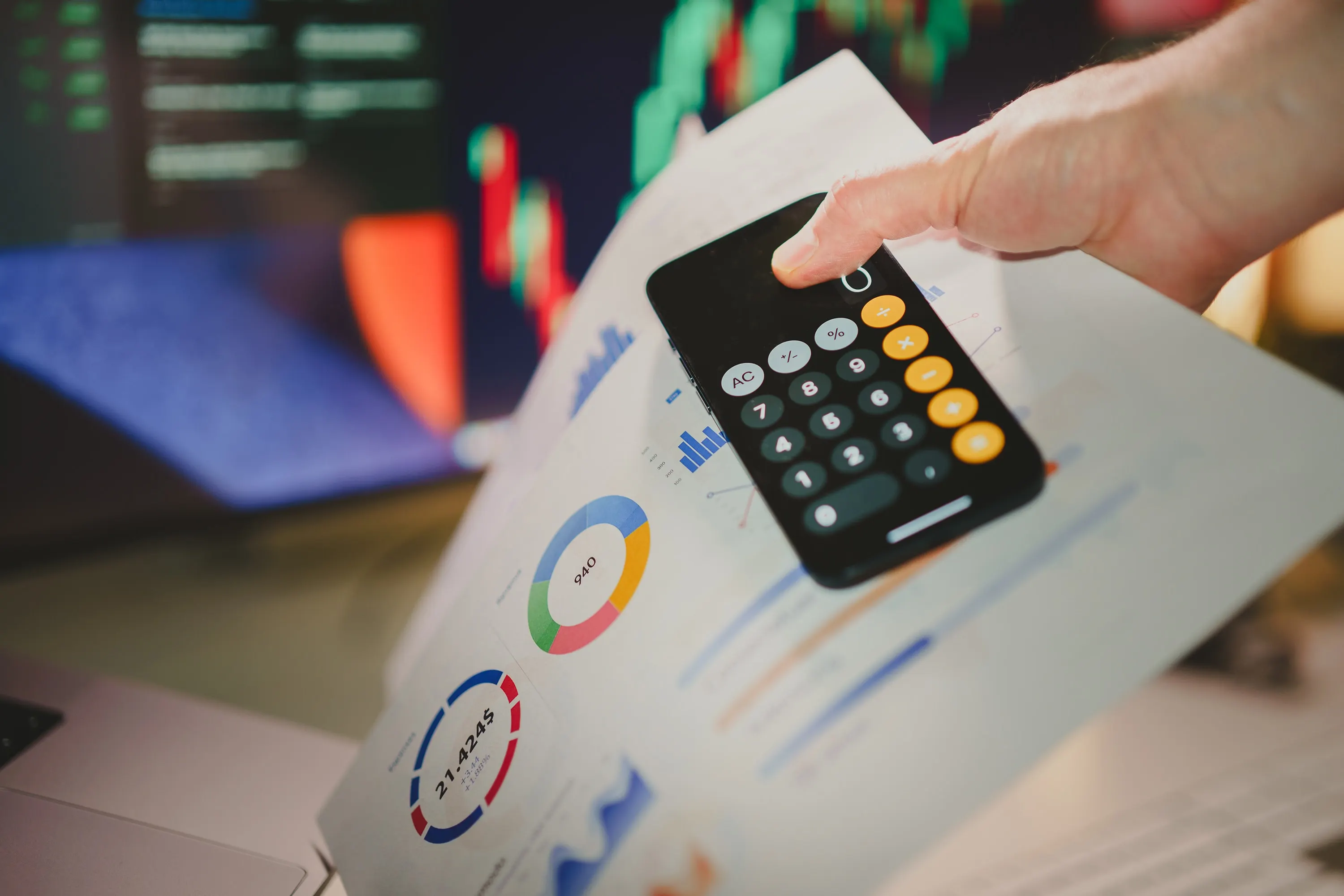 A hand using a smartphone calculator over financial charts.