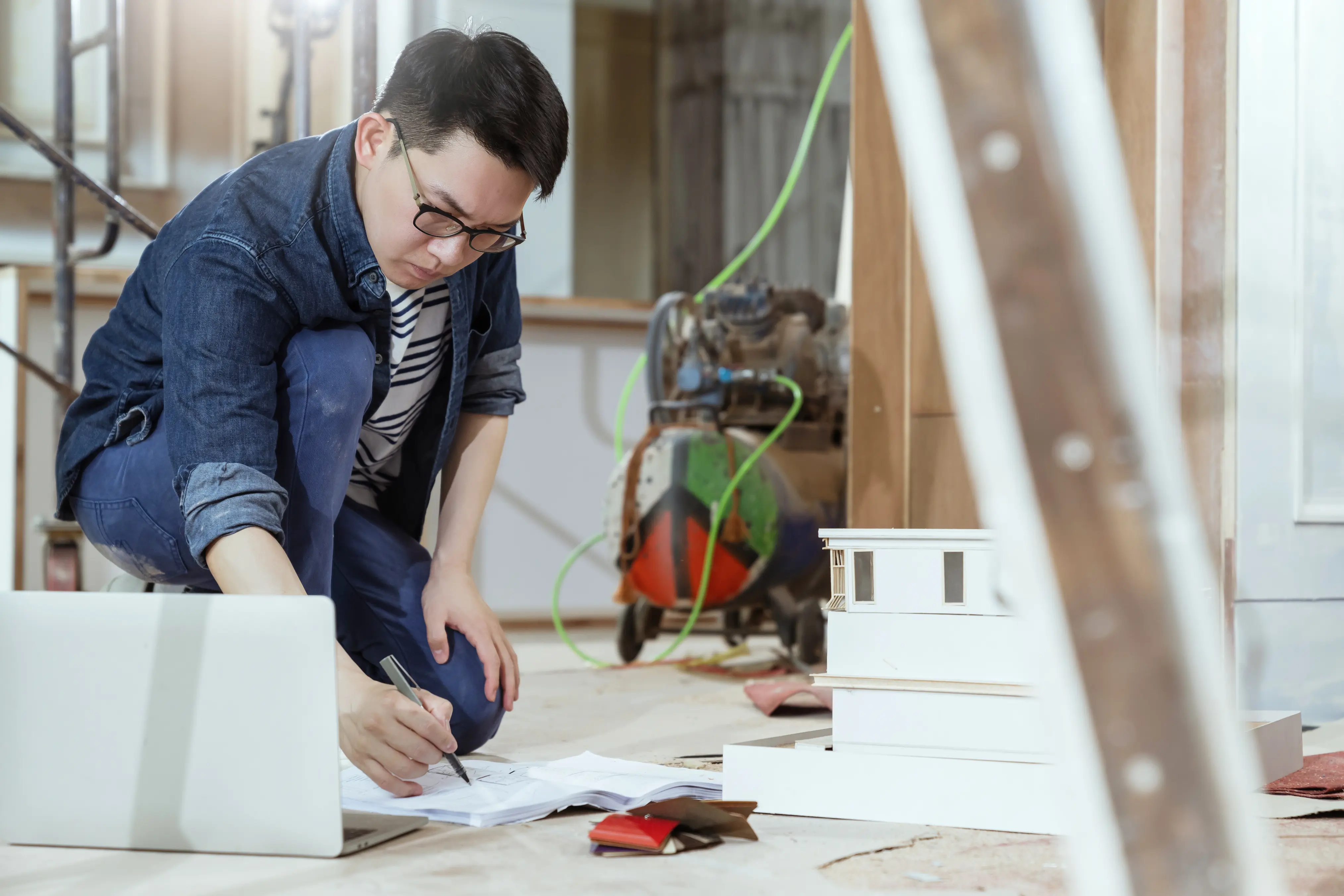 Carpenter working inside a building, writing on a notebook with a pen, while kneeling next to a laptop.