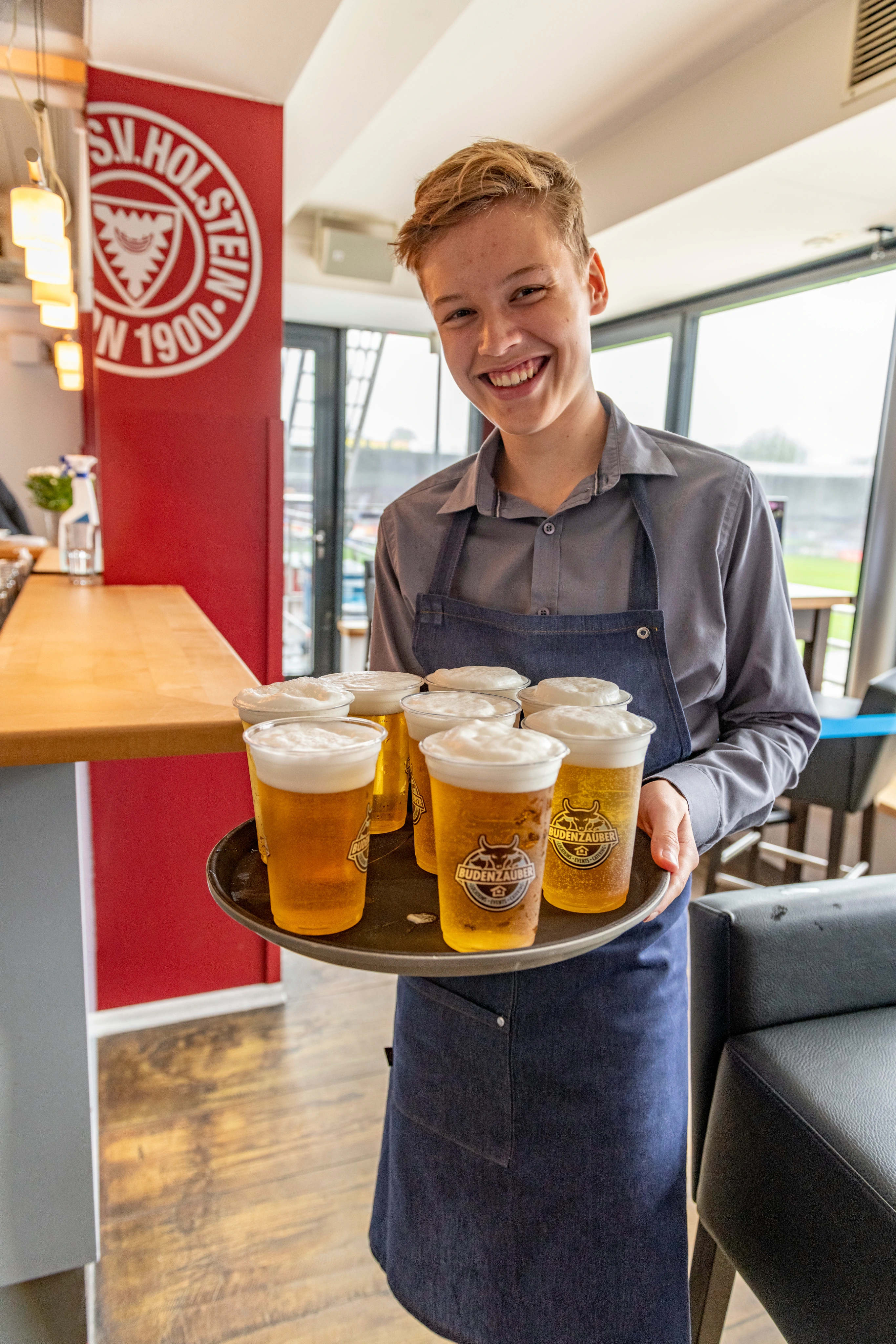 Junger Kellner trägt Tablett mit acht Bieren in Plastikbechern in einer Bar mit Holstein-Logo.