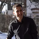 Junger Mann in schwarzer Jacke lächelt im Freien an einem sonnigen Wintertag.