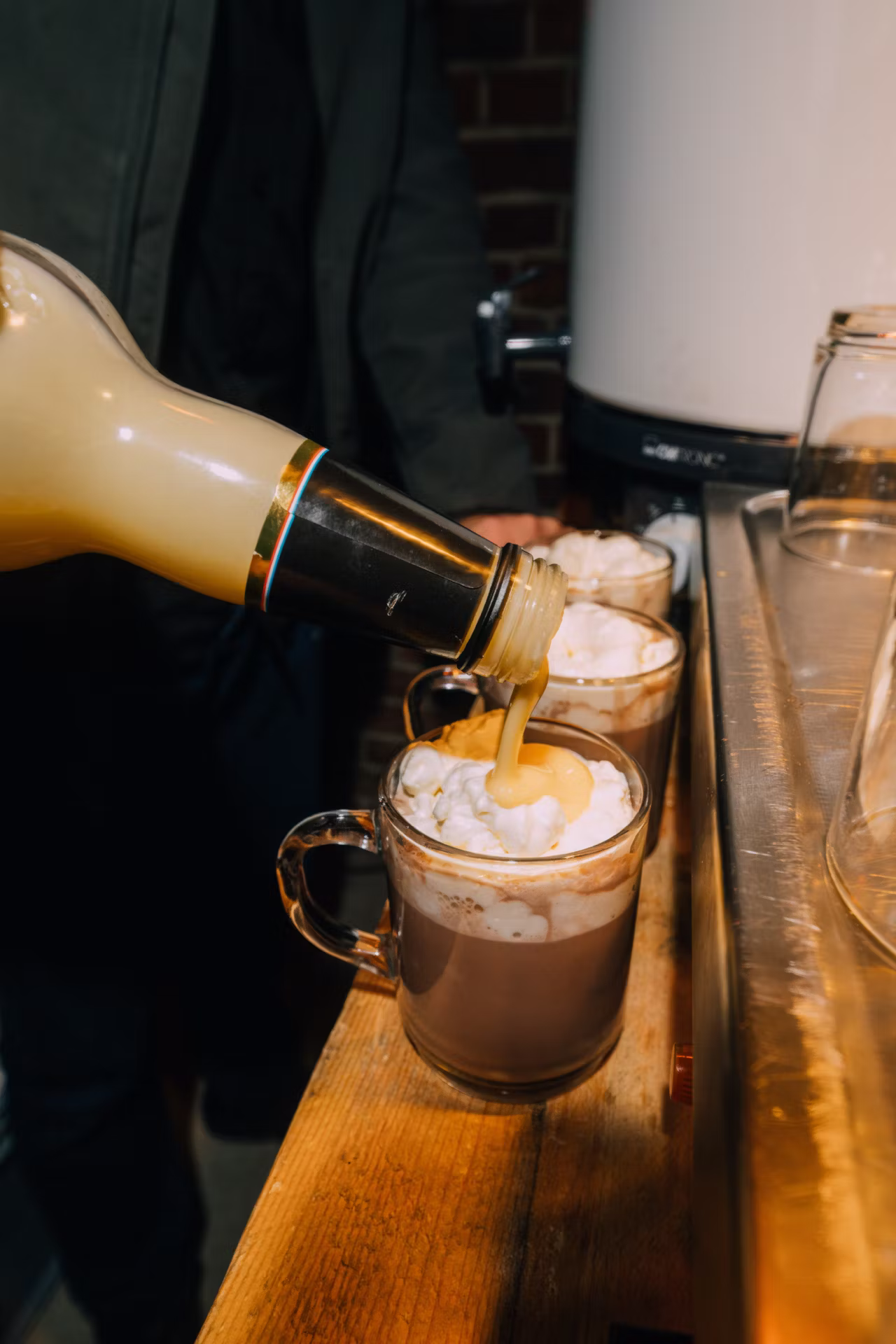 Eine Flasche gießt eine cremige Flüssigkeit auf heiße Schokolade mit Marshmallows in Glastassen.