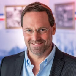 Mann mit Brille, hellblauem Hemd und dunklem Sakko lächelt frontal in einer Büro- oder Arbeitsumgebung.