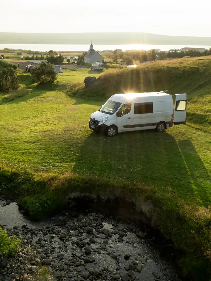 Ein weißes Wohnmobil, das auf sonnenbeschienenem Gras in der Nähe eines Baches geparkt ist, mit einer malerischen Landschaft und Wasser im Hintergrund.