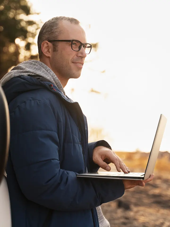 Mann mit Brille und blauer Jacke, der im Freien einen Laptop benutzt, lächelt und in die Ferne schaut.