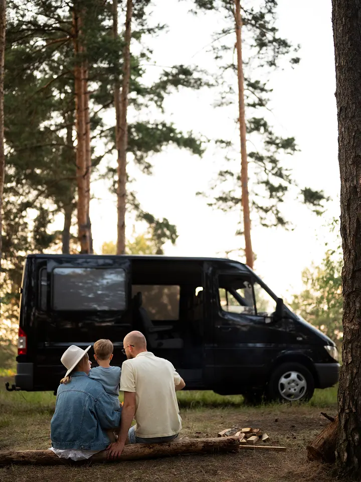 Rückenansicht einer Familie, die auf einem Baumstamm vor einem Schwarzen Campervan in einem Wald sitzt.