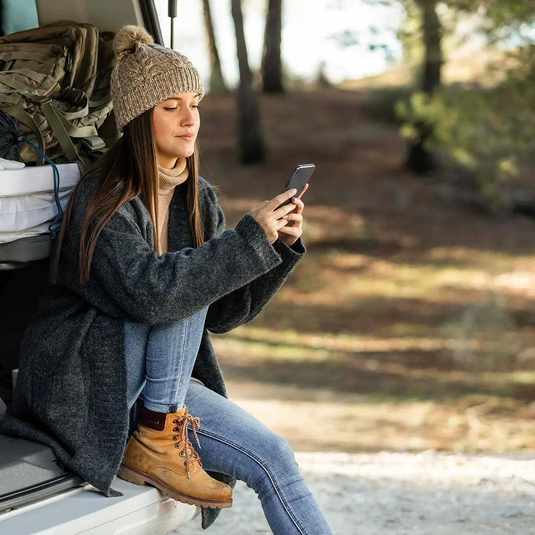 Frau mit Mütze und Stiefeln sitzt auf einem Campervan-Kofferraum im Wald und schaut auf ihr Telefon.