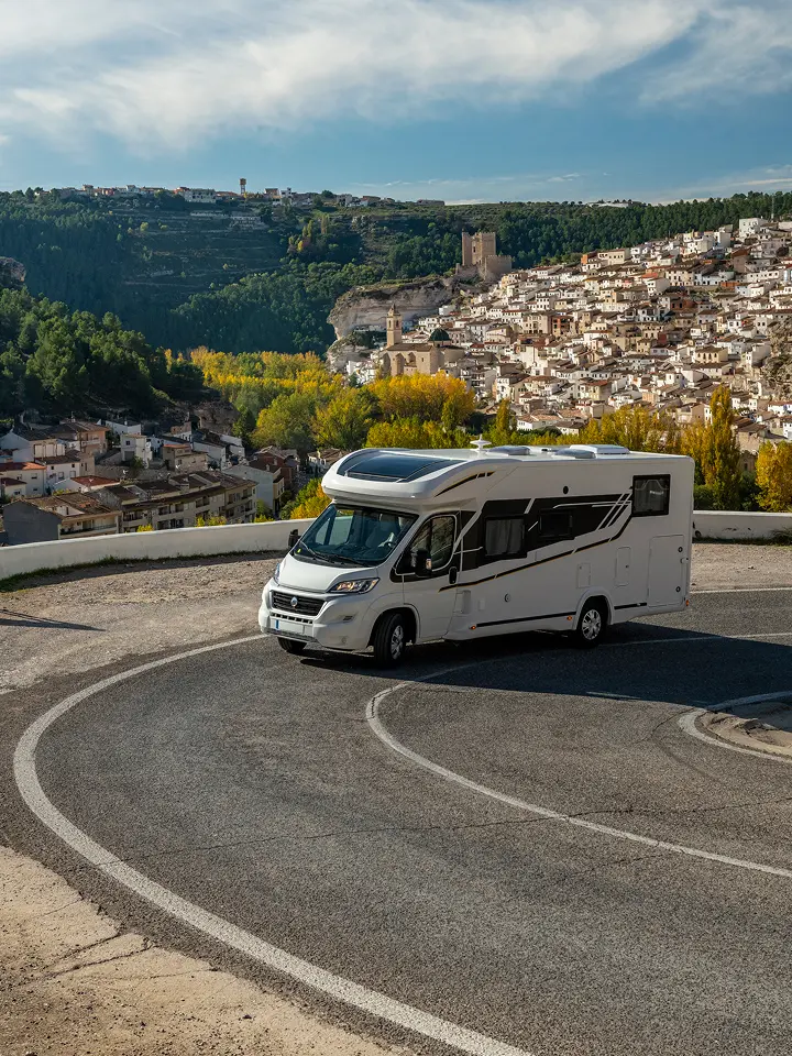 Ein weißes Wohnmobil fährt eine kurvenreiche Straße entlang und überblickt eine Stadt am Hang mit vielen Häusern und grünen Bäumen.