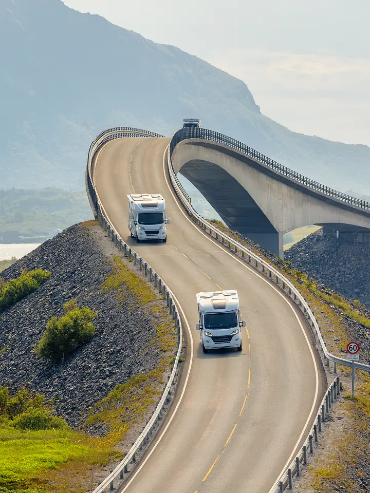 Zwei Wohnmobile fahren auf einer kurvenreichen, erhöhten Küstenstraße mit Bergen im Hintergrund.